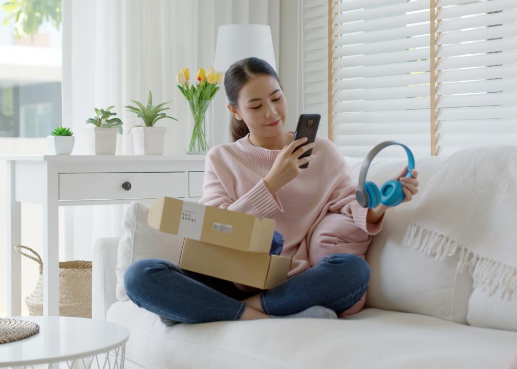 A woman is sitting on the couch with two boxes in her lap while taking a photo of her new headphones with her cell phone.
